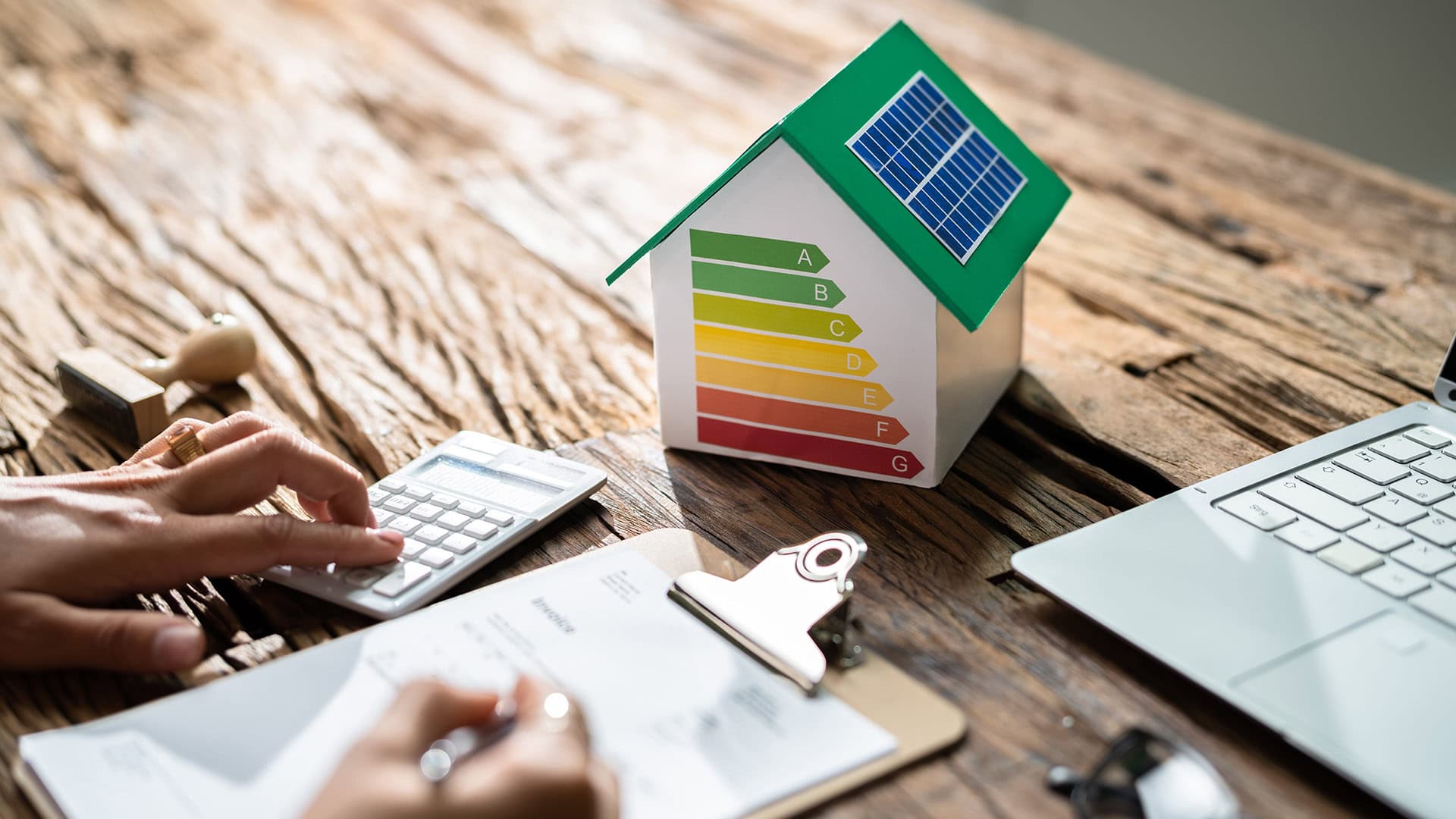 Detailný pohľad na model zeleného energeticky úsporného domu so solárnym panelom a energetickou triedou A, vedľa ruky používajúcej kalkulačku na výpočet úspory nákladov v zrekonštruovanom bytovom dome.